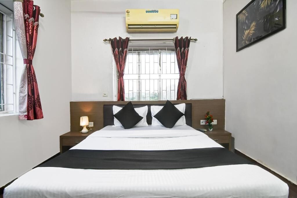 A hotel room with a bed, a nightstand, a lamp, a window with curtains, an air conditioner, and a wall-mounted picture.