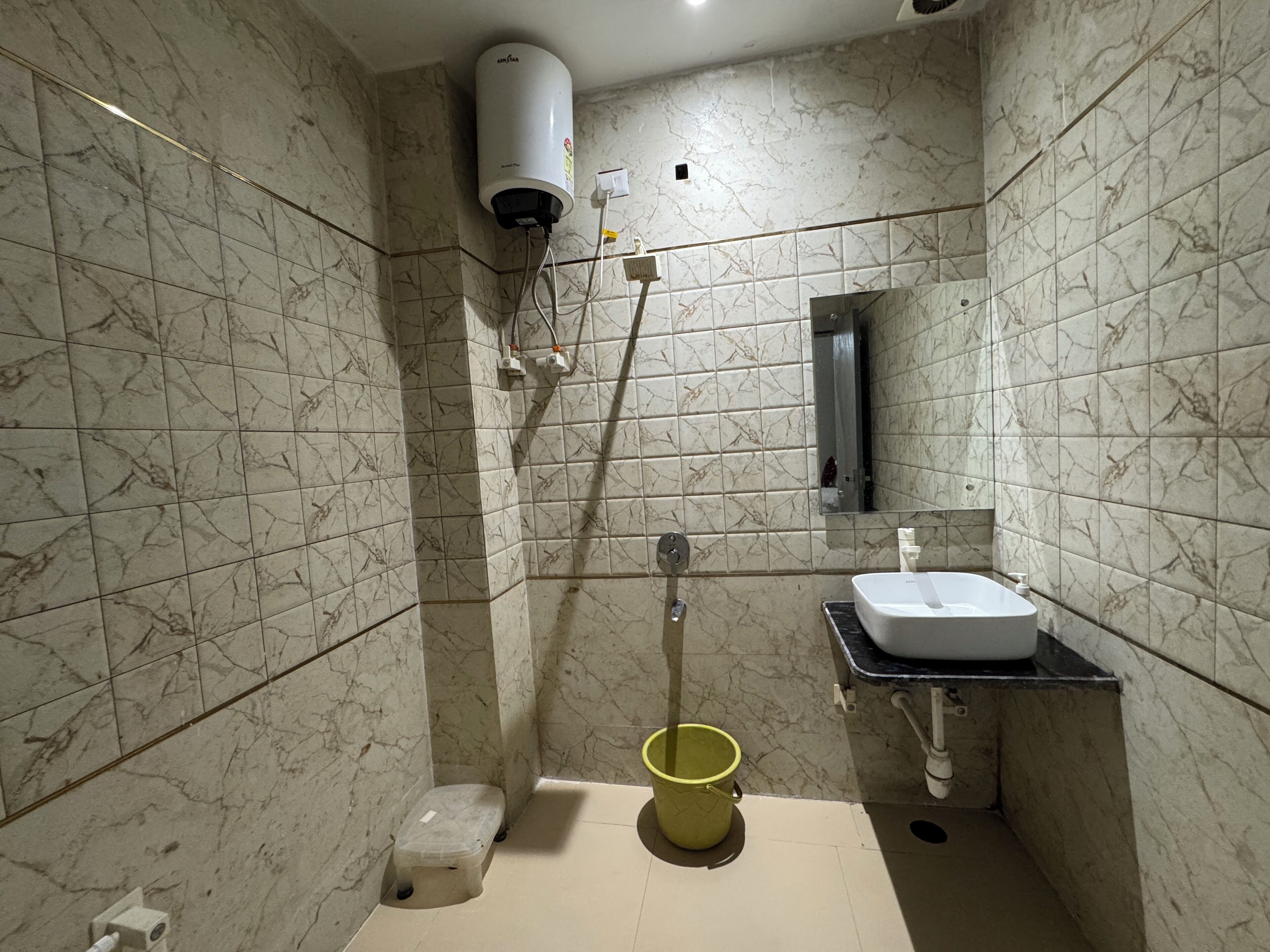 Ein Badezimmer mit Marmorfliesen, einem weißen Waschbecken, einem Spiegel, einem gelben Eimer und einem weißen Heizkörper.