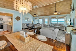A spacious living area with a large sofa, a dining table set with plates, and a view of the ocean through large windows. - Coconut Cottage at Cape San Blas (Gulf County)