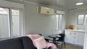 Ein gemütlicher, kompakter Wohnraum mit einem dunkelgrauen Sofa, einem kleinen runden Tisch und einer Küchenzeile mit weißen Schränken und einem Fenster.