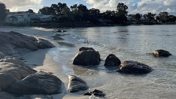 Ein Strand mit großen Felsen im Wasser, einem klaren Himmel und Häusern im Hintergrund.