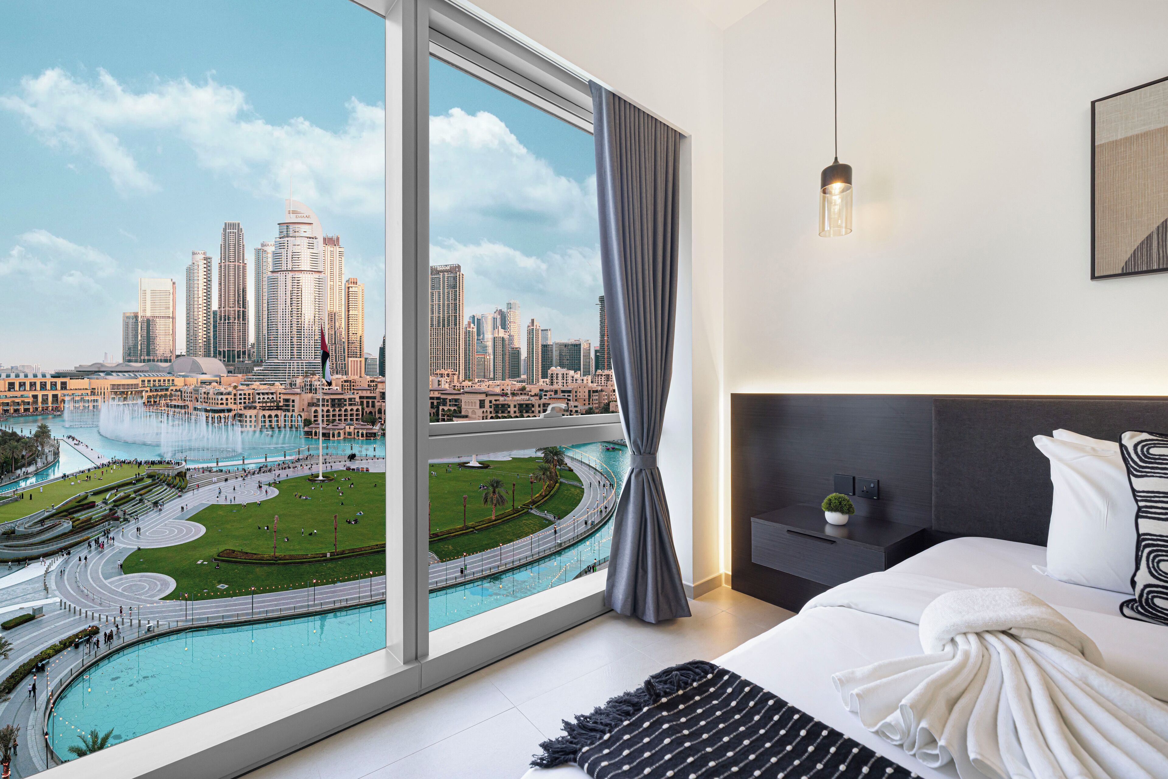 A modern hotel room with a large window offering a city view, a neatly made bed, a bedside table with a lamp, and a dark headboard.