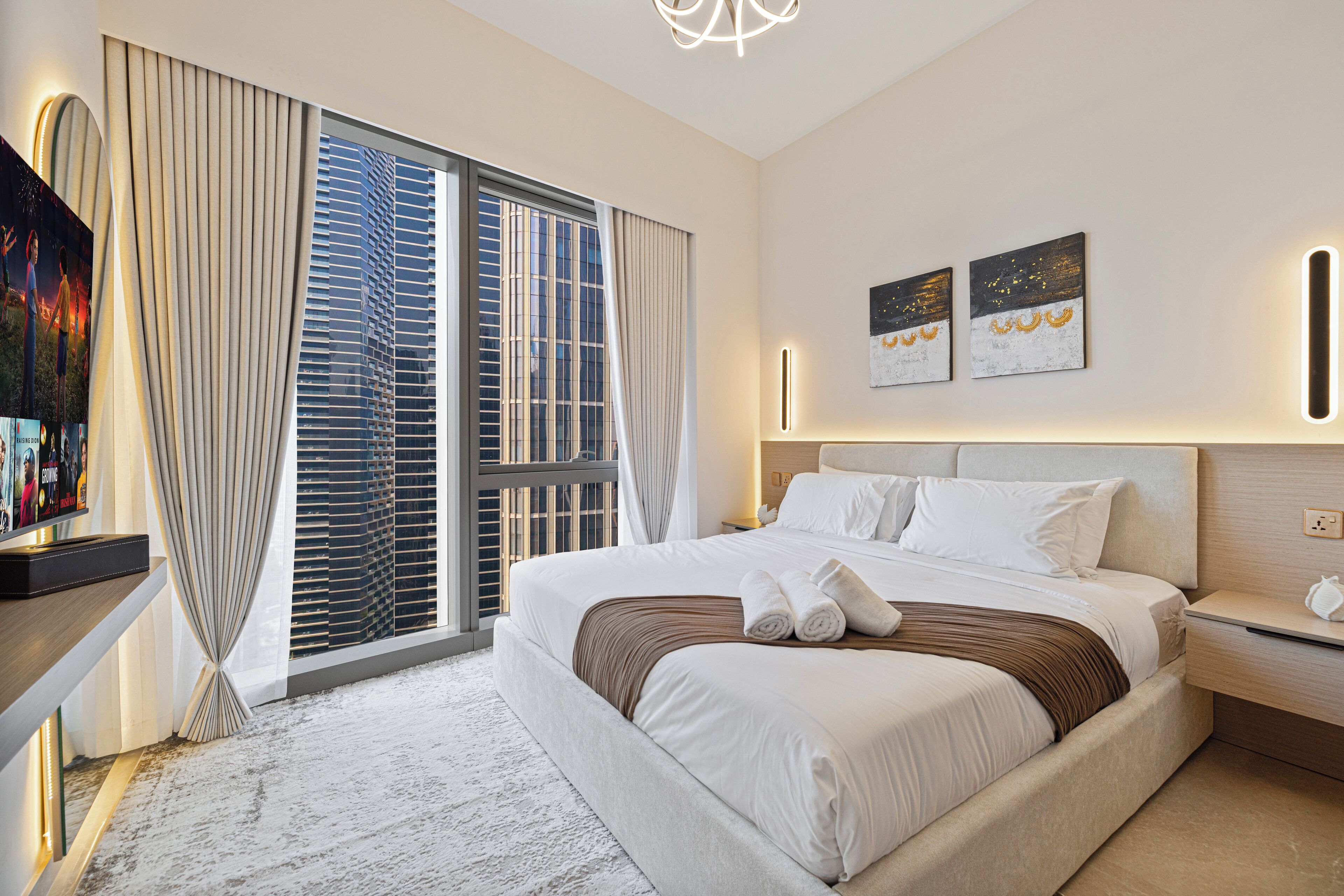 A modern hotel room with a large bed, a TV on a stand, and a view of a cityscape through the window.