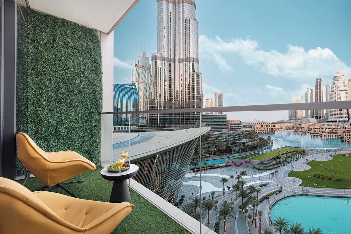 A modern hotel room with a balcony overlooking a cityscape, a pool, and a landscaped area.