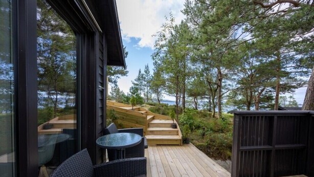A wooden deck with outdoor furniture, a glass table, and a view of trees and a body of water. - Nordica Resort Florø (Kinn)