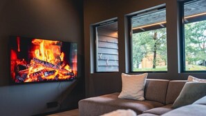A modern living room with a flat-screen TV displaying a fire, a brown sofa, and large windows with a view of greenery. - Nordica Resort Florø (Kinn)