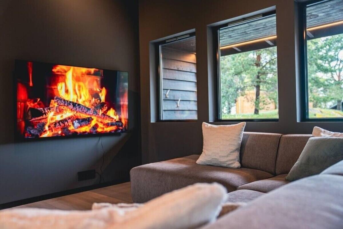 Una sala de estar moderna con un televisor de pantalla plana mostrando un fuego, un sofá marrón y ventanas amplias con vistas a la vegetación.