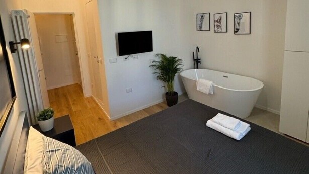 Un dormitorio moderno con una cama grande, una lámpara colgante negra, un televisor de pantalla plana, una bañera blanca y una planta en maceta.