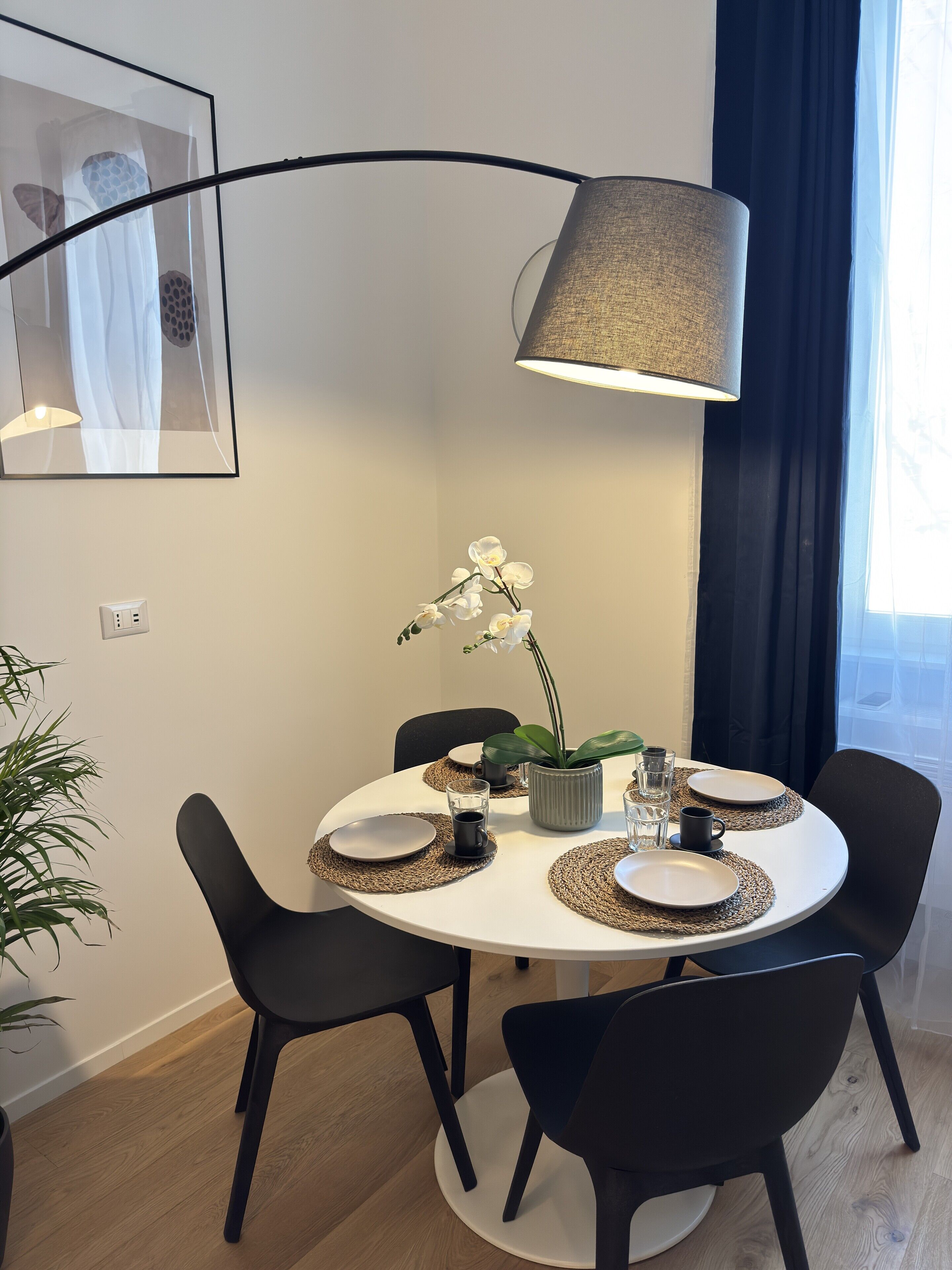A modern dining area with a round table, black chairs, woven placemats, and a potted orchid.