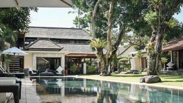 Un área de piscina con sillones de descanso y sombrillas, rodeada de vegetación tropical y un edificio con techo de paja.
