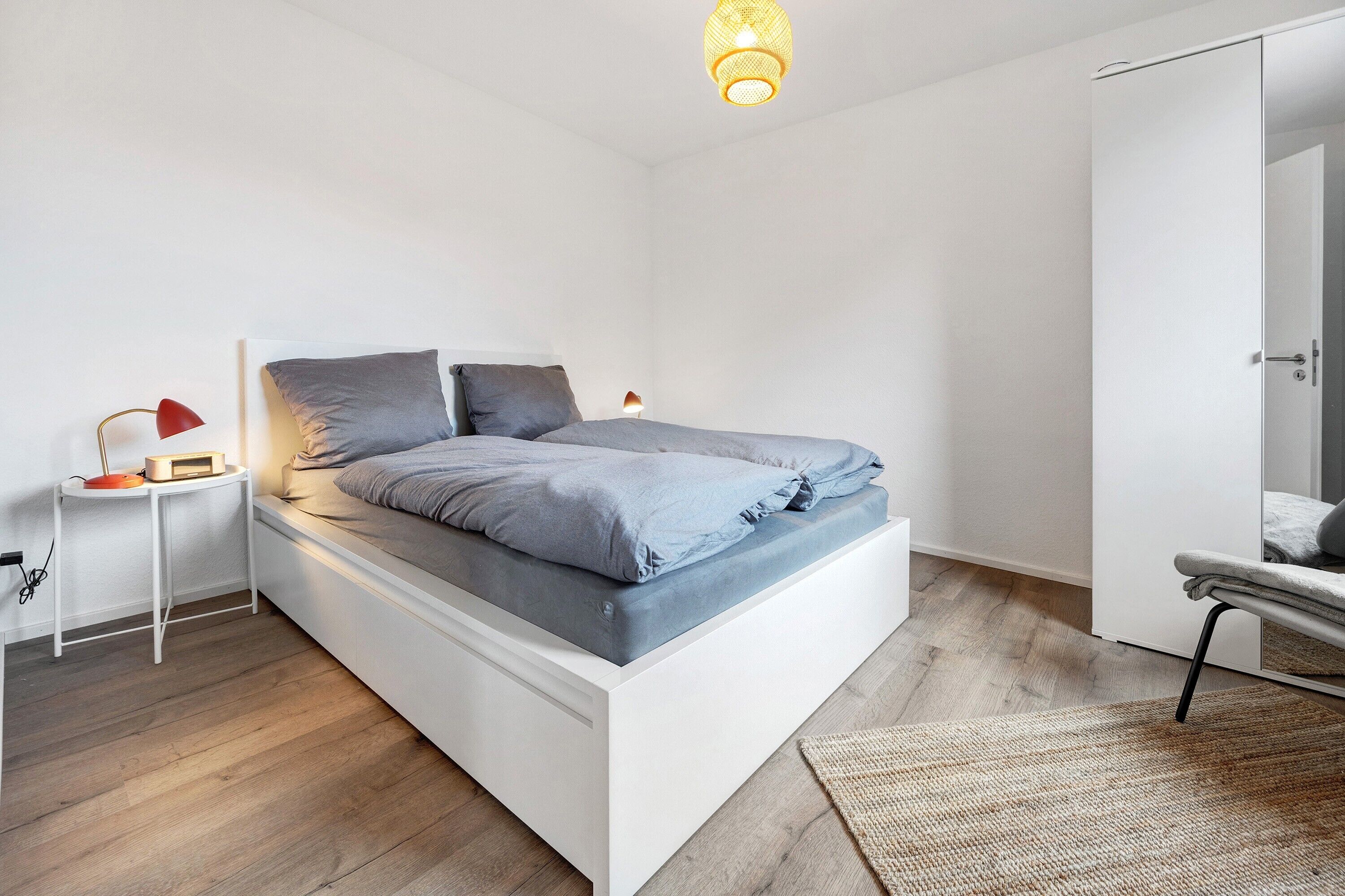 Ein modernes Schlafzimmer mit einem weißen Bett, einem Nachttisch mit Lampe und Holzboden.