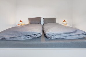 Un lit double avec une literie grise, deux tables de chevet avec des lampes et un mur blanc uni.