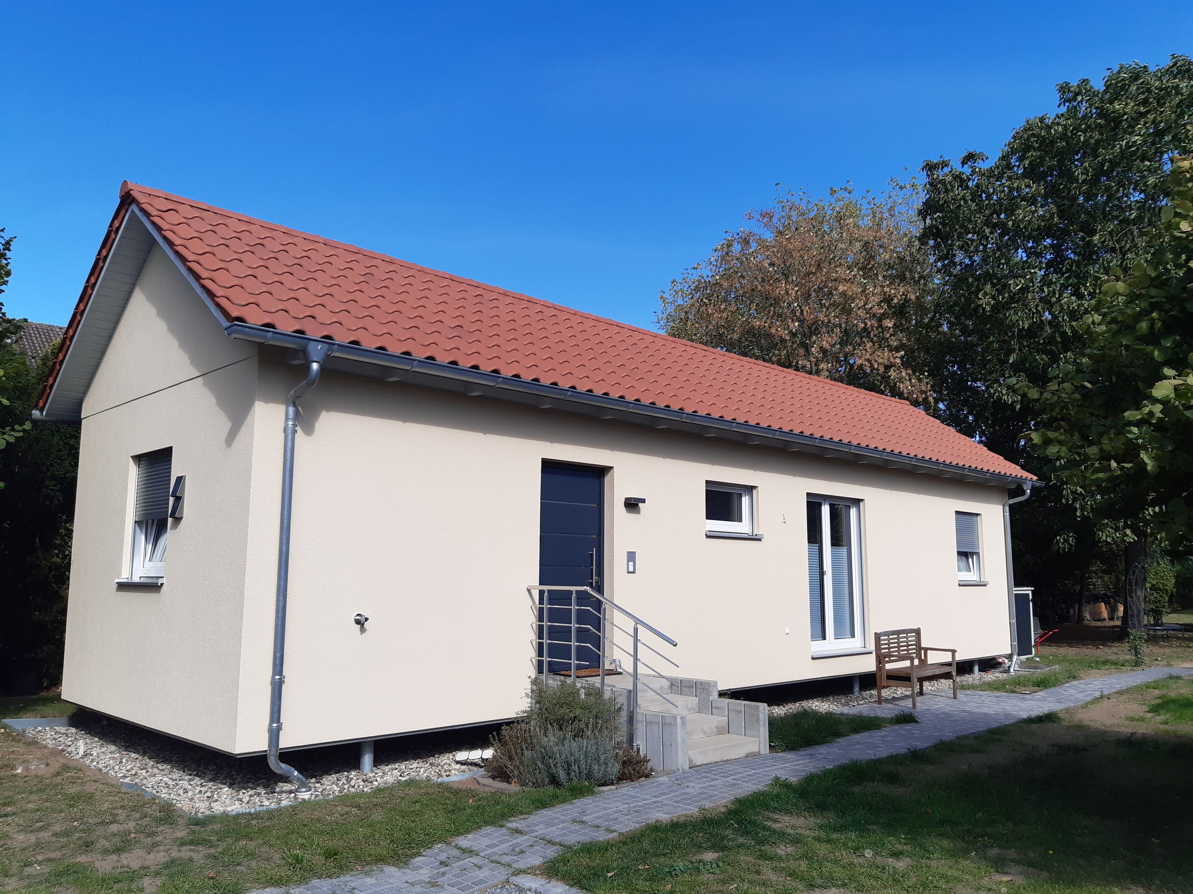Ein eingeschossiges Haus mit rotem Ziegeldach, einer Haustür und einer kleinen Veranda mit einer Bank.