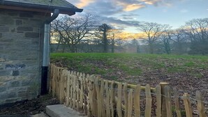 A stone building with a metal gutter, a wooden fence, and a grassy field with trees in the background. - Historic Gardener’s House in Haltinne (Gesves)