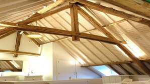 A bedroom with a sloped ceiling, wooden beams, a bed, and a bathroom with a vanity. - Historic Gardener’s House in Haltinne (Gesves)