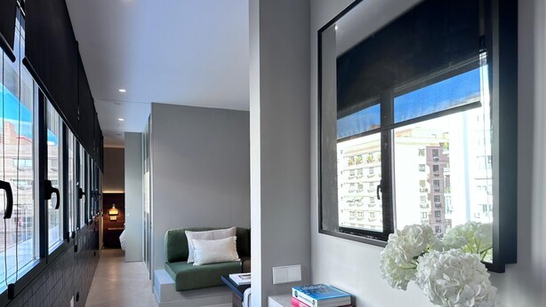A modern hotel room with a long hallway, a green sofa, a small table with books and a candle, and a window with a city view. - Edmond Vibes Almagro Suites (Madrid)