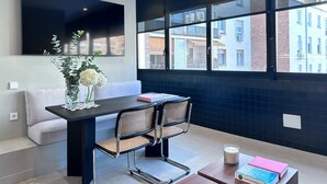 A modern room with a large window, a flat-screen TV mounted on the wall, a dining table with two chairs, a sofa, and a vase with flowers. - Edmond Vibes Almagro Suites (Madrid)