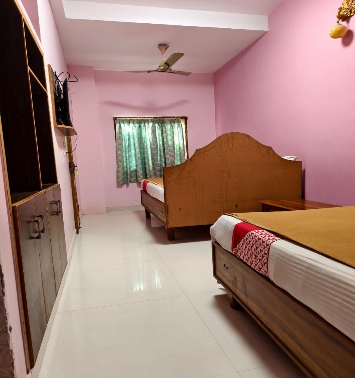 Une chambre avec deux lits, un ventilateur de plafond, une fenêtre avec des rideaux et un téléviseur posé sur un meuble.
