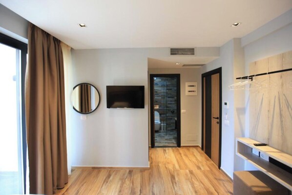 A modern hotel room with a wooden floor, a flat-screen TV mounted on the wall, a mirror, and a wooden panel with a hanging rack.