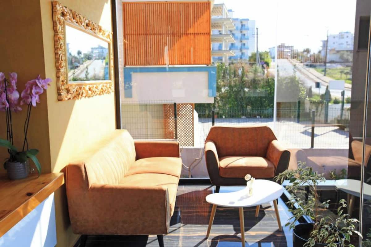 A modern living room with a sofa, armchair, and coffee table, featuring a large window with a view of buildings and greenery.