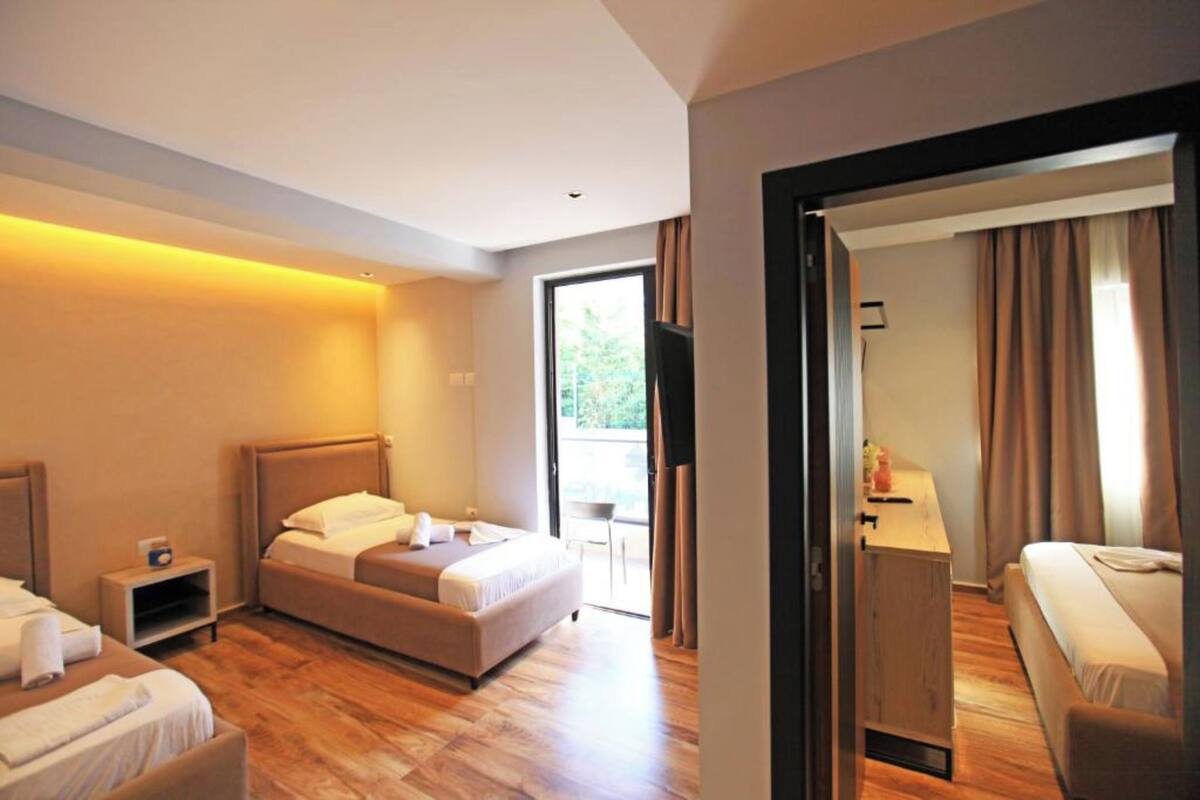 A hotel room with a bed, a desk, and a chair. There is a window with curtains and a view of greenery outside.