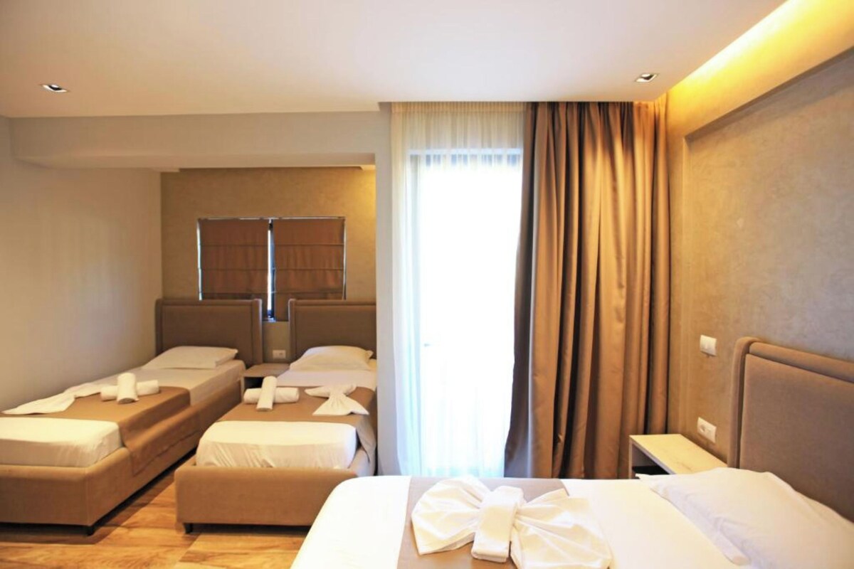 A hotel room with two single beds, a larger bed, a window with curtains, and a mirror.
