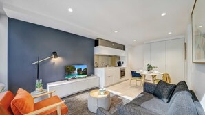 A modern living room with a sofa, a coffee table, and a TV. There is a kitchen area with cabinets and a dining table.