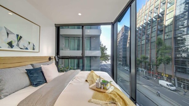 A modern hotel room with a large bed, a city view, and a framed abstract painting on the wall.