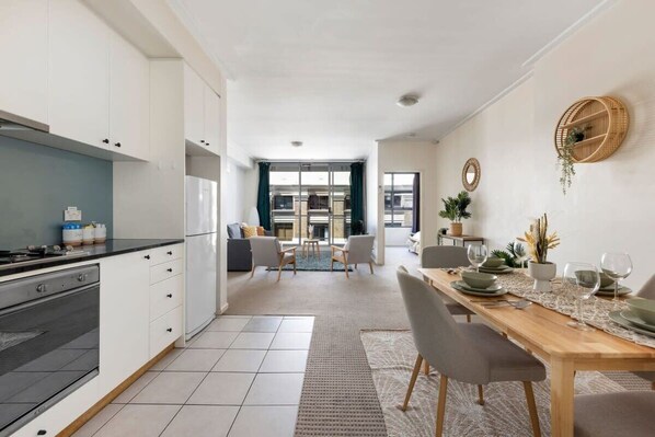 A modern kitchen with white cabinets, a black countertop, and a dining area with a wooden table set for four. - A Plush & Comfy Apt Right Next to Darling Harbour (Sydney)