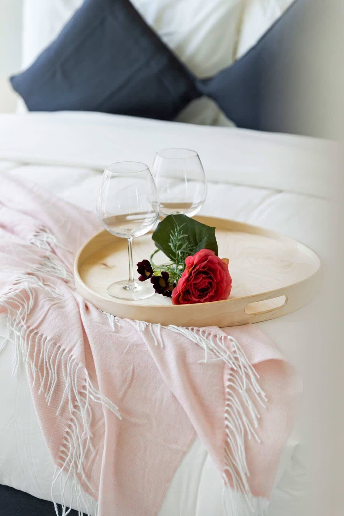 A bed with a white and blue pillow, a pink blanket, and a tray with two glasses of water and a red rose.