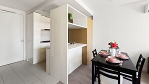 Eine moderne Küche mit weißen Schränken, ein Essbereich mit einem schwarzen Tisch für vier Personen und eine kleine Pflanze auf einem Regal.