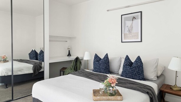 A modern hotel room with a large bed, a bedside table with a lamp, a mirror, and a framed picture on the wall. - Cozy 2BR Apt, Steps From Melb Central With Parking (Melbourne)