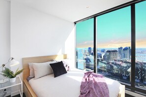A modern bedroom with a large city view, a bed with a wooden headboard, a bedside table with a lamp and a plant, and a large window. - Stylish CBD Apartment With Stunning Harbour Views (Melbourne)