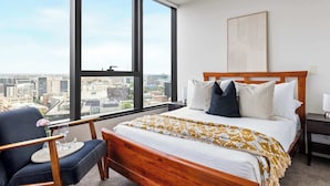 A hotel room with a large window, a bed with a wooden headboard, a chair with a cushion, and a view of the cityscape. - A Modern & Cozy 2BR Apartment With City Views (Melbourne)