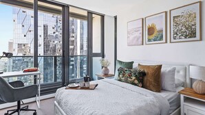 A modern bedroom with a large window, a bed with white bedding, a desk with a chair, and artwork on the wall.