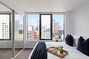 Une chambre d’hôtel avec un grand lit, un plateau avec un vase de fleurs et une vue sur la ville à travers de grandes fenêtres.