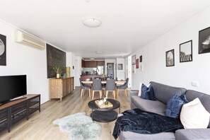 A modern living room with a flat-screen TV, a grey sofa, and a dining area with a wooden table and chairs.