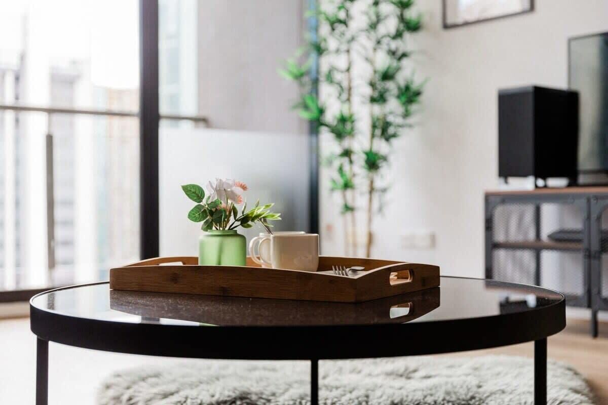 A round glass table with a wooden tray holding a green vase with flowers, a white mug, and a spoon.