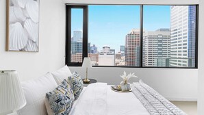 A modern hotel room with a large window offering a city view, a neatly made bed, and a decorative vase on the nightstand. - Modern 2BR Apt Next to Southern Cross Free Parking (Melbourne)