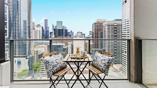 A balcony with a table and two chairs, overlooking a cityscape with high-rise buildings. - Modern 2BR Apt Next to Southern Cross Free Parking (Melbourne)