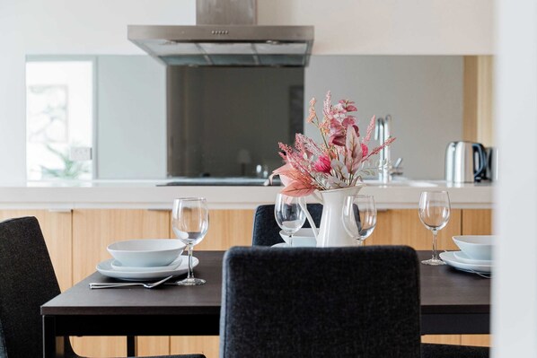 Un comedor con una mesa dispuesta para dos personas, un florero con flores y una cocina moderna al fondo.
