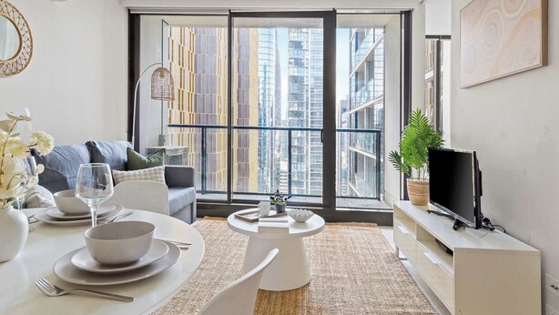 Un salon moderne avec un coin repas, de grandes fenêtres et un téléviseur posé sur une console blanche.