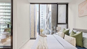 Une chambre moderne avec un grand lit, une vue sur de hauts immeubles et une porte-fenêtre coulissante donnant sur un balcon.