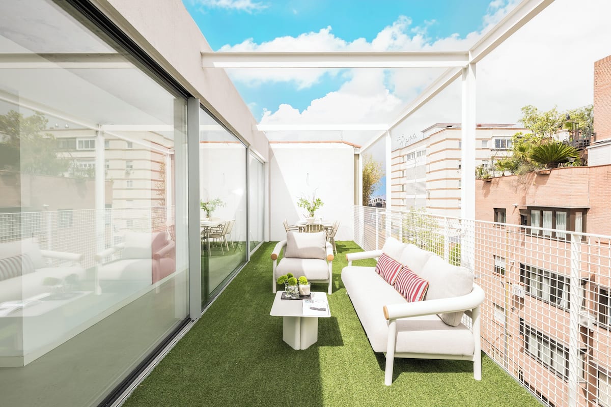 A modern balcony with a glass wall, a white sofa, a small table, and a green artificial turf floor.