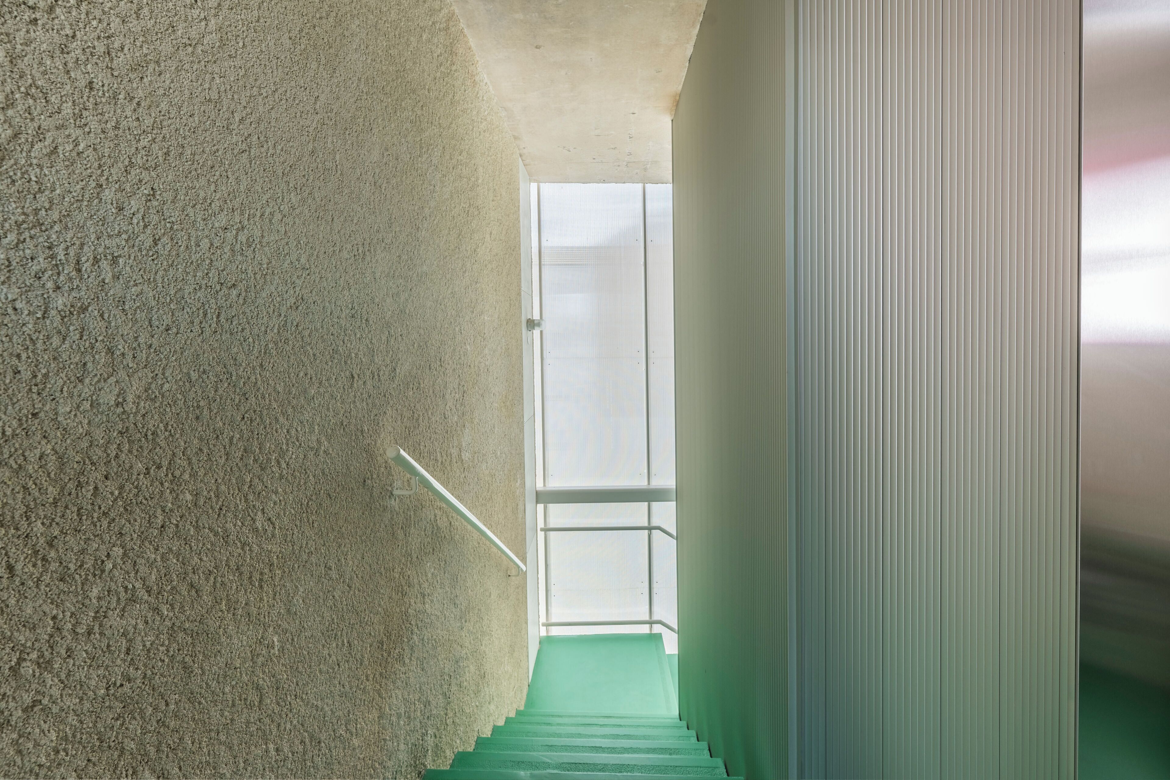 Una escalera con un muro con textura a un lado y un muro liso al otro, que conduce a un espacio interior luminoso.