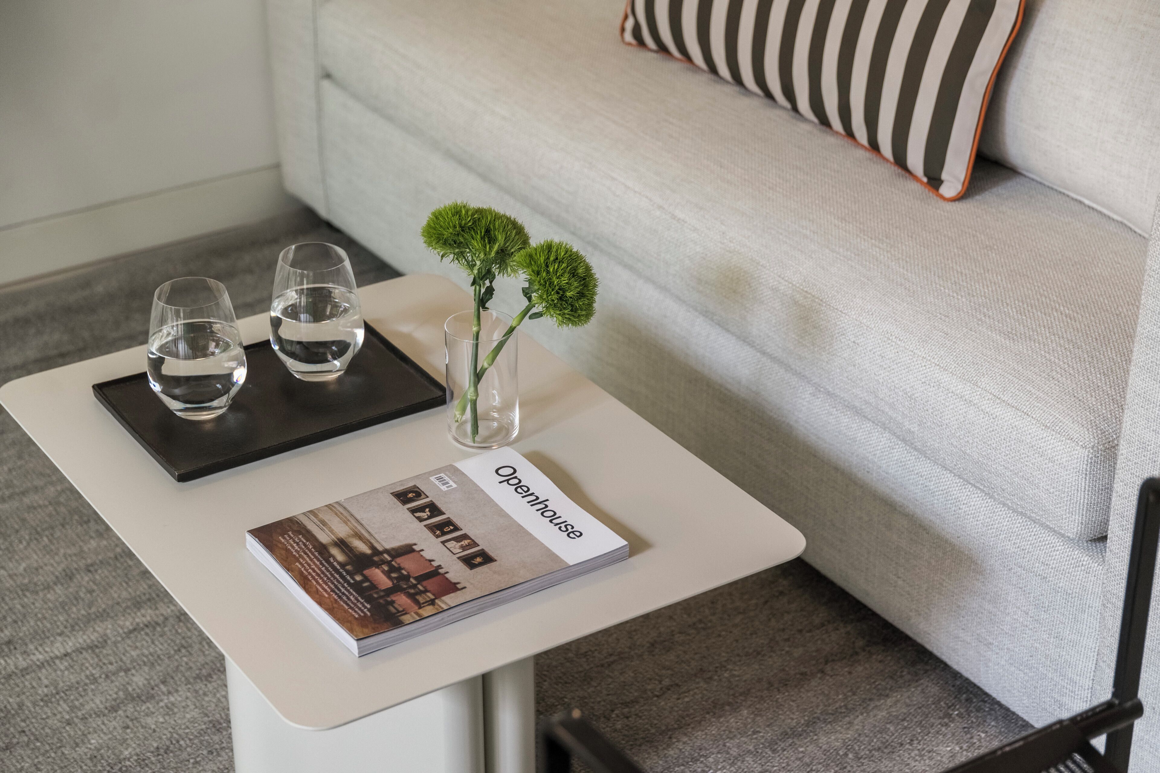Una mesa blanca con dos vasos de agua, un jarrón pequeño con flores verdes y una revista titulada 'Openhouse'.