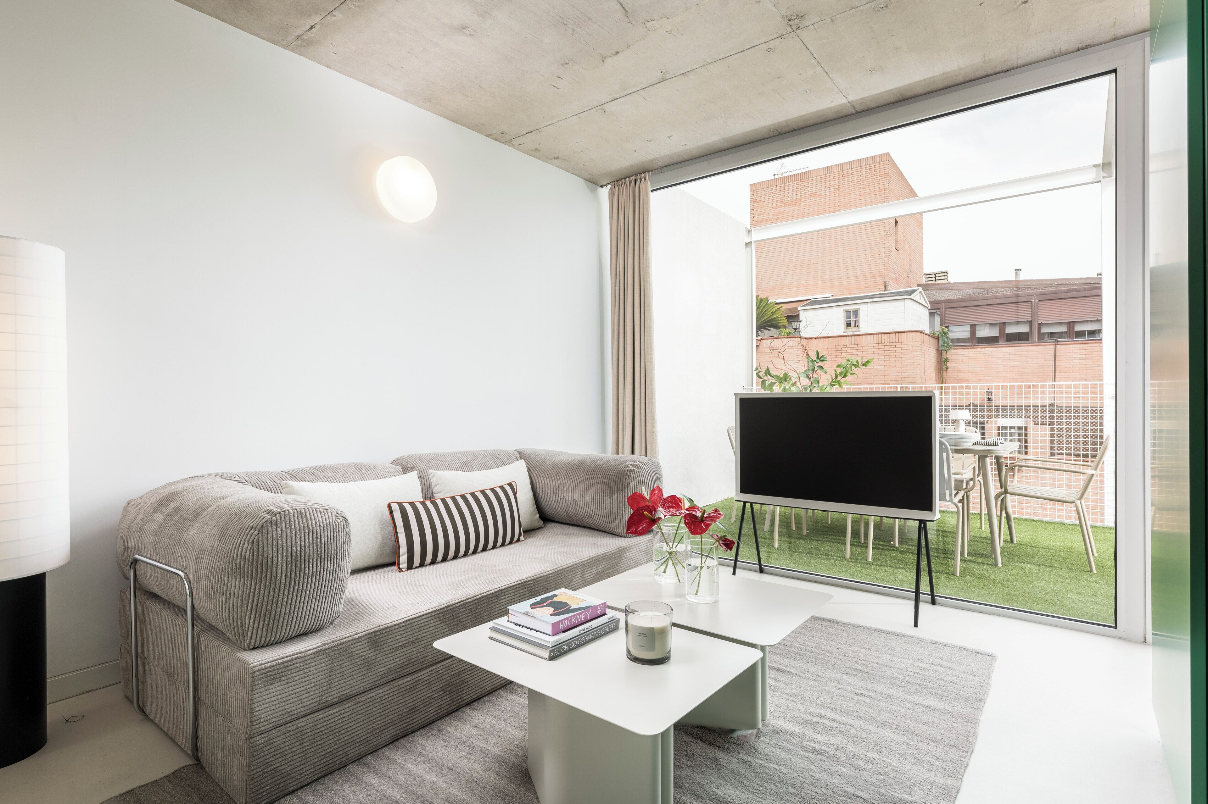 Una sala moderna con un sofá, un televisor de pantalla plana, una mesa de centro y vistas a un edificio de ladrillo a través de ventanas grandes.