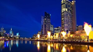 A nighttime cityscape with illuminated buildings reflecting on a calm body of water. - A Cozy 2BR Apt on Collins Near Flinders Station (Melbourne)