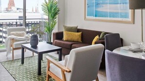 A modern living room with a sofa, armchairs, a coffee table, and a dining table set for two. A large window offers a view of a cityscape with a prominent building. - A Cozy 2BR Apt on Collins Near Flinders Station (Melbourne)
