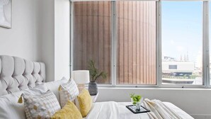 A bedroom with a large window, a bed with patterned pillows, a bedside table with a lamp, and a view of buildings outside. - A Cozy 2BR Apt on Collins Near Flinders Station (Melbourne)